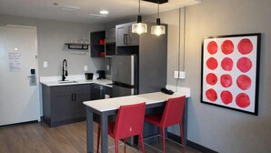 A modern kitchen with grey cabinets, a stainless steel refrigerator, and a white countertop. There are two red chairs at a white table.