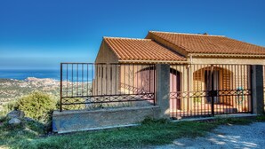 Un edificio pequeño con techo de tejas y portón metálico, rodeado de vegetación y con vistas a un paisaje costero.