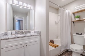 Een badkamer met een wit wastafel, spiegel en toilet, een douche met een witte douchegordijn, en een houten plank met handdoeken.