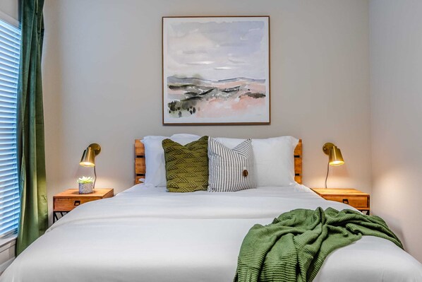 Een net opgemaakt bed met witte lakens, een groene plaid en twee bedlampjes.