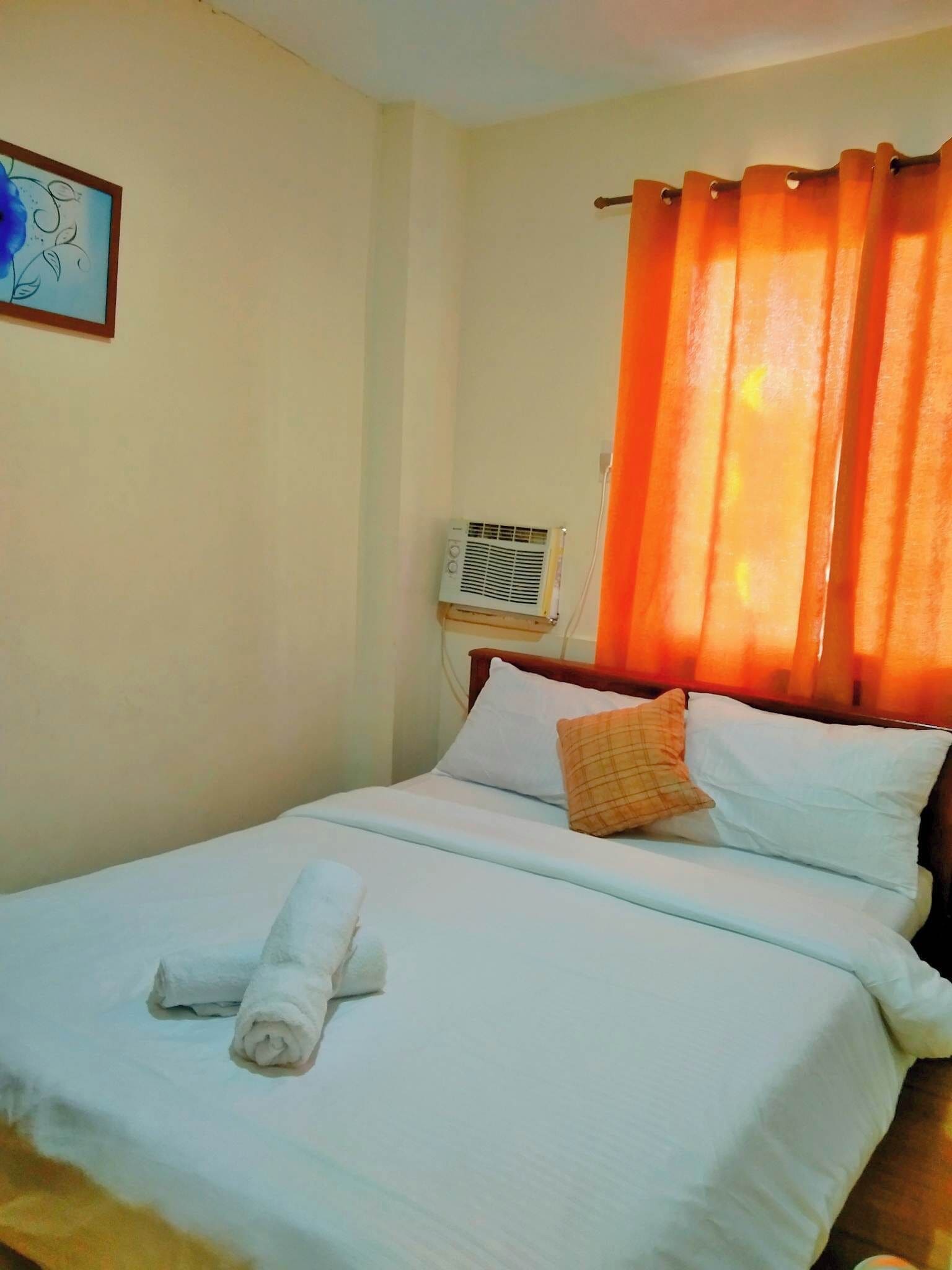 A neatly made bed with white linens and a rolled towel, a wall-mounted air conditioner, and a single framed picture.