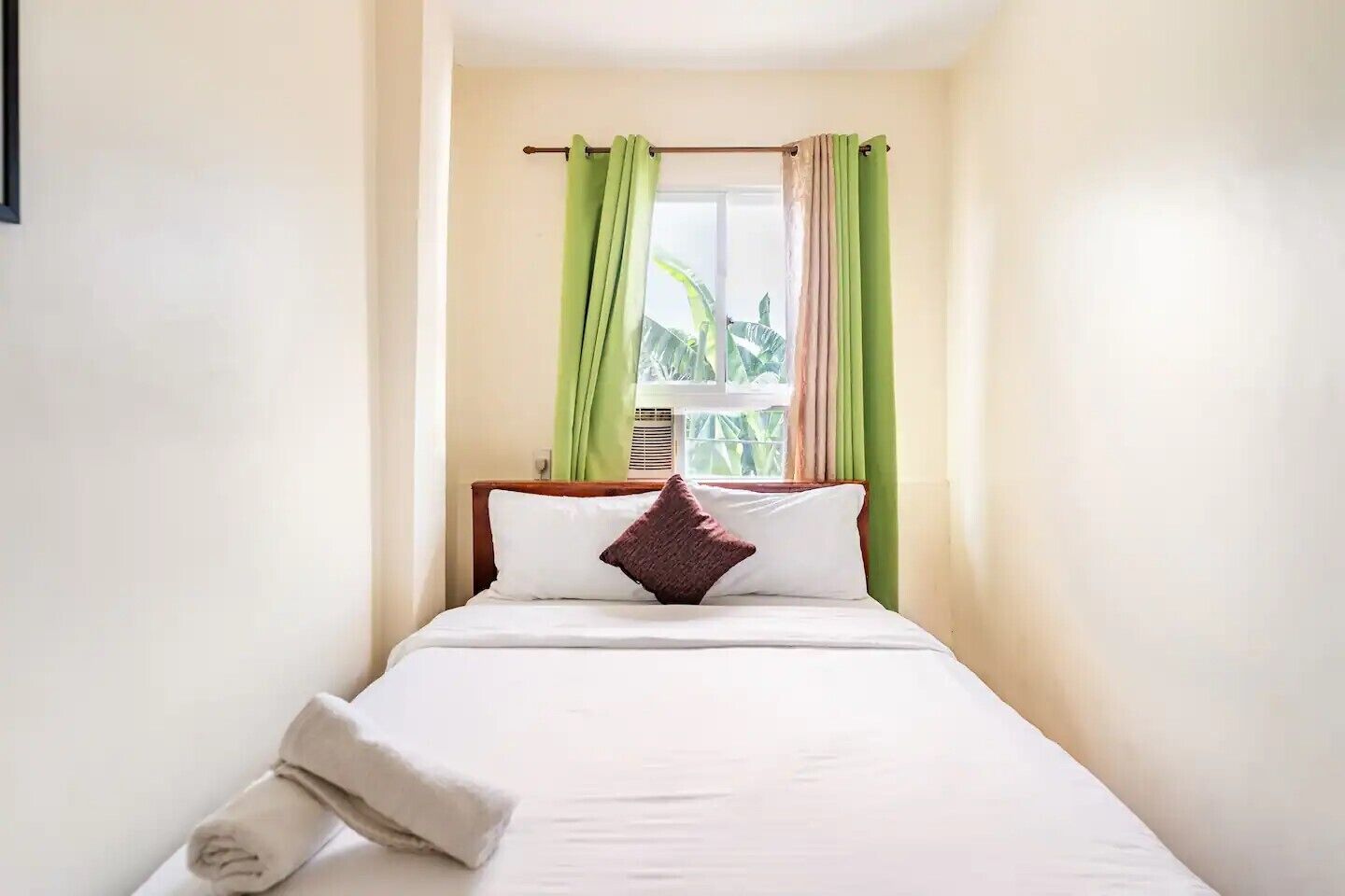Ein ordentlich bezogenes Bett mit weißen Leinen und einem einzelnen braunen Kopfkissen, ein grün-beiger Vorhang und ein Fenster mit Blick ins Grüne.