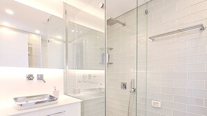 Un bagno moderno con cabina doccia in vetro, pareti piastrellate di bianco, un mobile lavabo bianco con specchio e un piccolo cestino.
