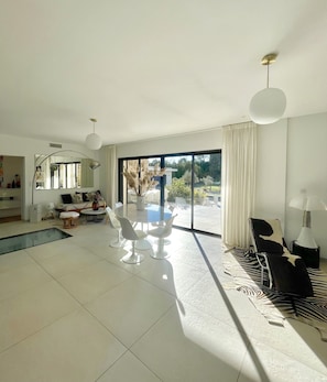 A spacious living room with a large sliding glass door leading to a balcony, a round dining table with chairs, a sofa, and a large mirror.