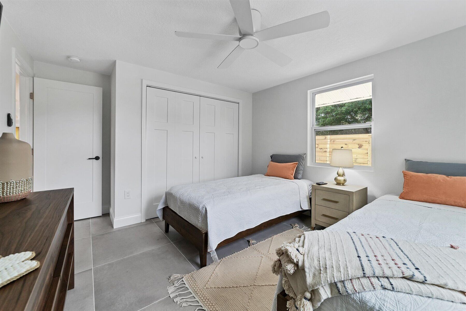 A bedroom with two beds, a ceiling fan, a window with a view, and a bedside table with a lamp.