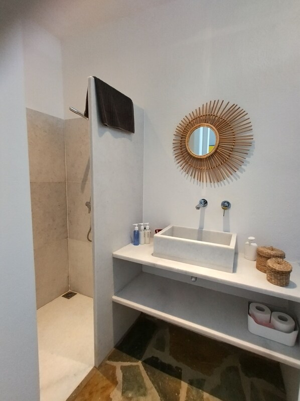 A bathroom with a shower, a sink with two faucets, a mirror with a sunburst frame, and shelves with toiletries.