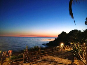 Vista costera al atardecer con un paseo de madera que conduce a la playa, rodeado de vegetación.