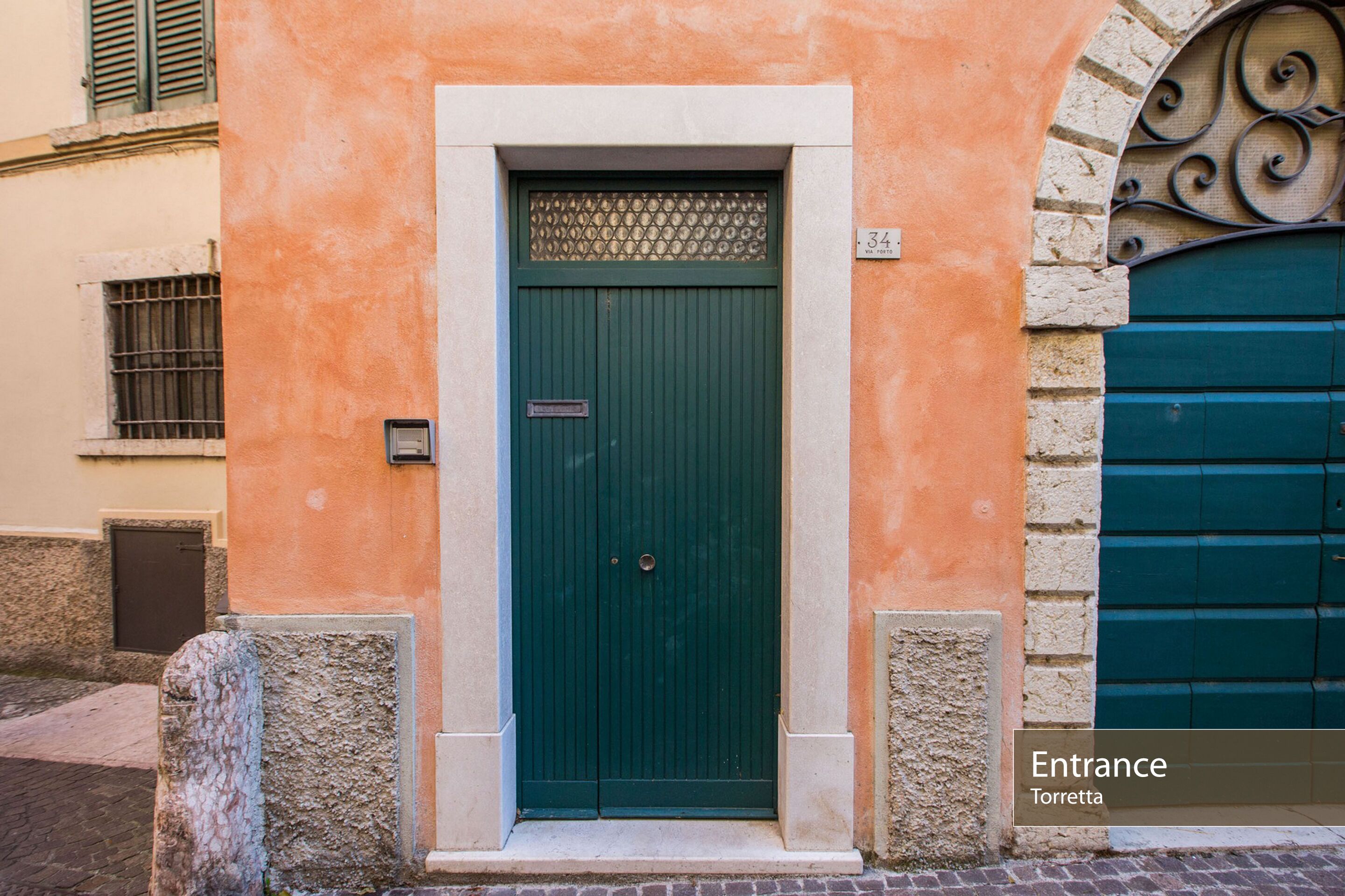Una porta verde con un cartello sopra, il numero '34' sul muro e un segnale che indica 'Ingresso Torretta'.