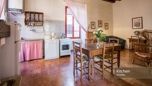 Una cocina con estufa blanca, un lavamanos y una mesa de comedor de madera con sillas. Hay una ventana con cortina roja y un ventilador de techo.