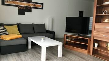 Ein modernes Wohnzimmer mit einem grauen Sofa, einem weißen Couchtisch, einem Fernseher auf einem Holzständer und einem Bücherregal.