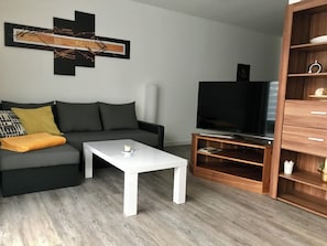Ein modernes Wohnzimmer mit einem grauen Sofa, einem weißen Couchtisch, einem Fernseher auf einem Holzständer und einem Bücherregal.