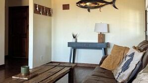 A living room with a wooden coffee table, a sofa with decorative pillows, a wall-mounted fish sculpture, a small table with a lamp, and a door leading to another room.