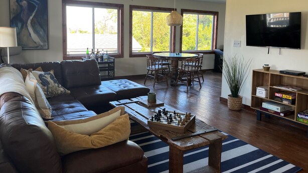 A living room with a brown leather couch, a wooden coffee table with chess, a dining table with chairs, a TV, and large windows with a view of trees.