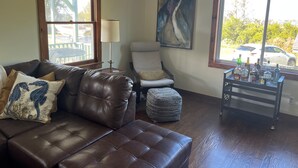 A living room with a brown leather sofa, a white armchair, a round ottoman, a painting, and a small table with bottles.