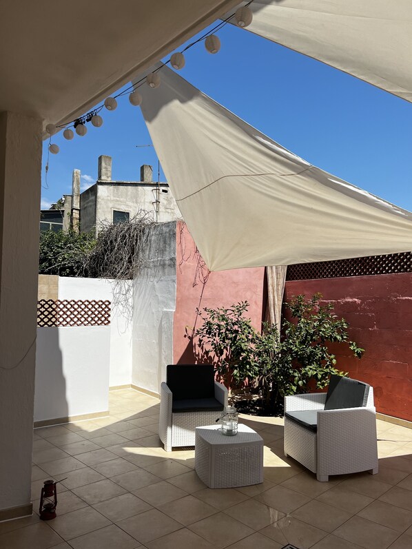 A covered outdoor seating area with two white wicker chairs and a small table, a red wall, and a white fence.