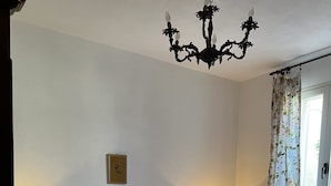 Ein Schlafzimmer mit einem großen Bett, zwei Nachttischen mit Lampen, einer Kronleuchter, einem Fenster mit floralem Vorhang und einem Wandbild.