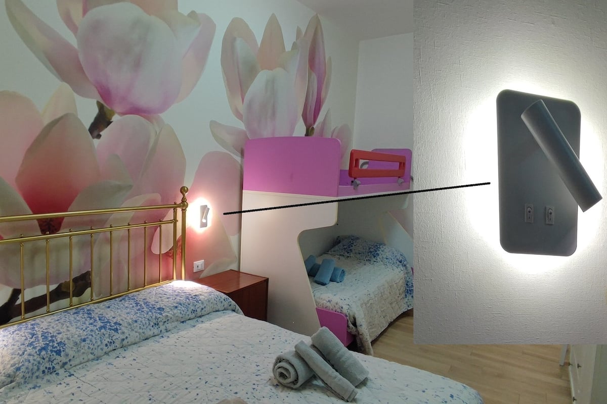 A bedroom with a bed, a nightstand, a wall-mounted lamp, a small table, and a wall-mounted phone.