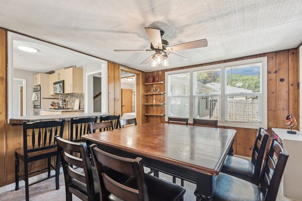 A dining area with a large wooden table and chairs, a ceiling fan, and a view of the kitchen and outside through large windows.