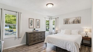 A bedroom with a large bed, a dresser, and a nightstand with a lamp. There are two windows with curtains, a ceiling light, and a wall-mounted painting. - Affordable & Spacious home near Minneapolis (Minneapolis)