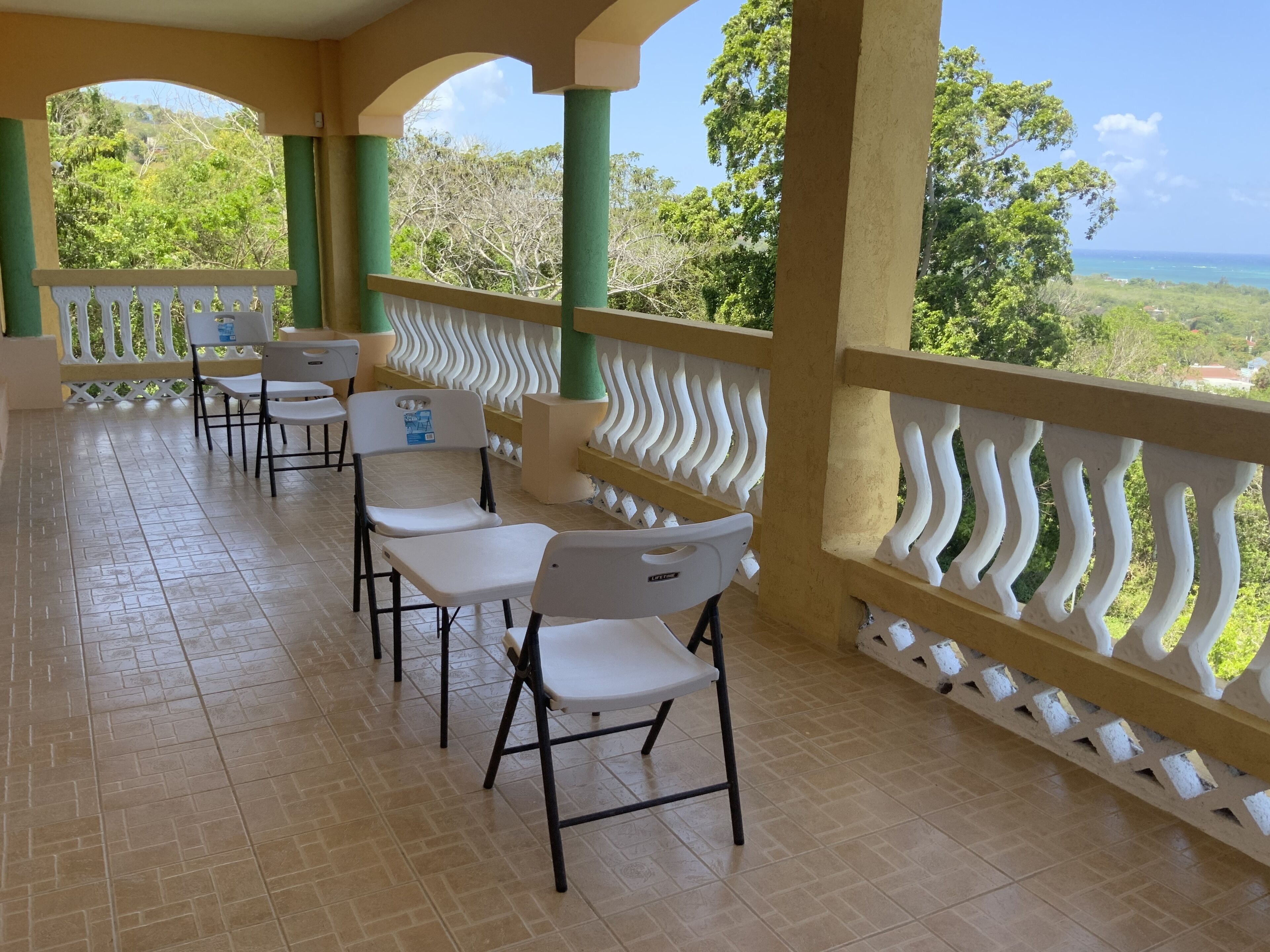 Een overdekt terras met witte stoelen en tafels, groene pilaren en uitzicht op zee en groen.