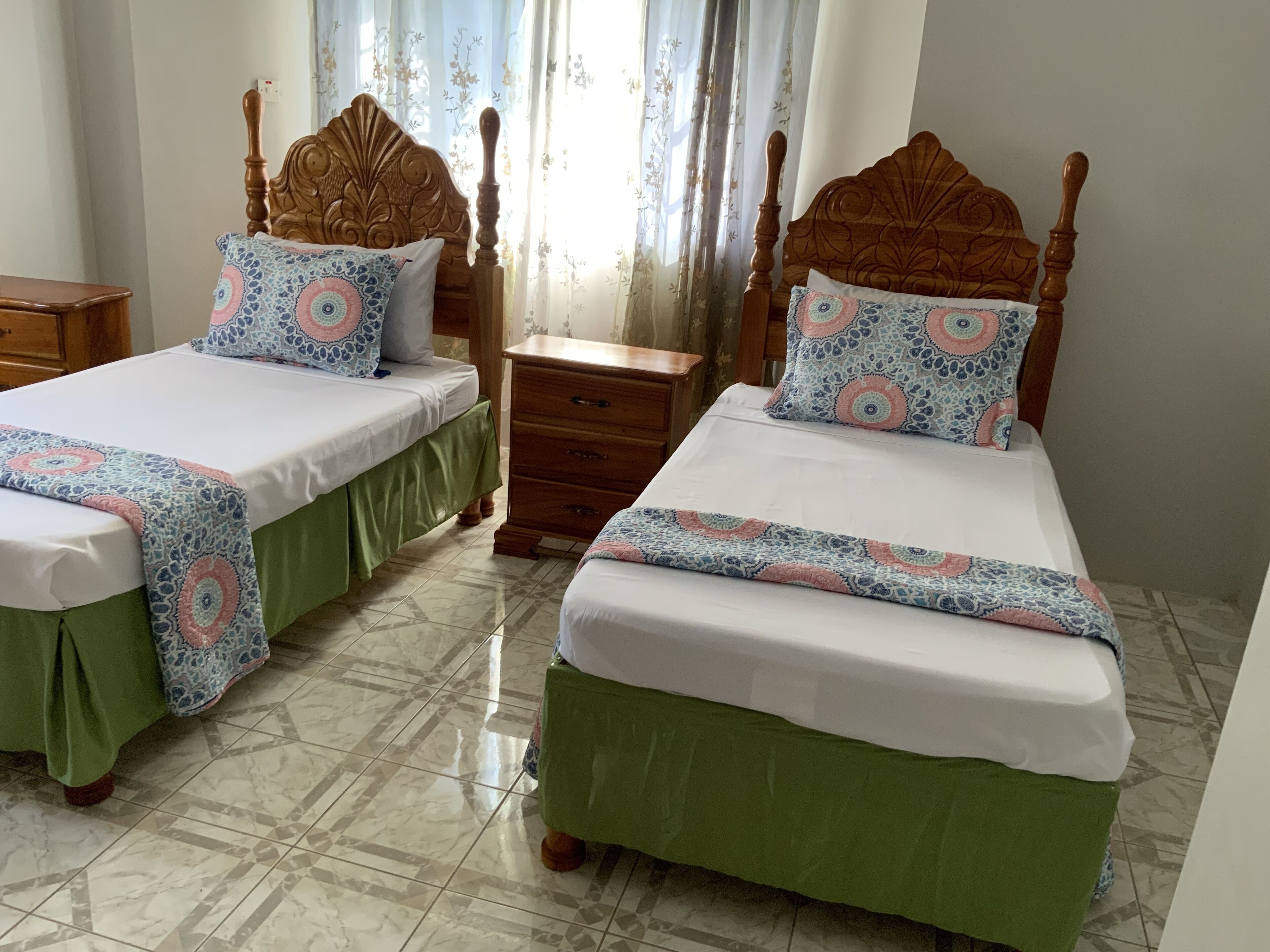 A room with two wooden beds, each with a green skirt and a patterned bedspread. There is a wooden nightstand between the beds, and a small wooden table to the left.
