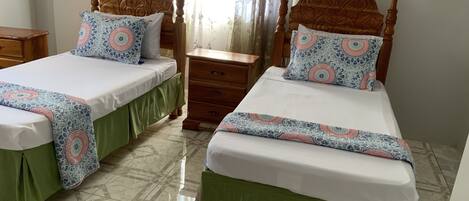 A room with two wooden beds, each with a green skirt and a patterned bedspread. There is a wooden nightstand between the beds, and a small wooden table to the left.