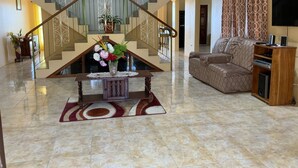 Uma sala de estar ampla com teto alto, escada com grades ornamentadas, um sofá grande, uma mesa de centro com um arranjo floral e uma televisão sobre um suporte de madeira.