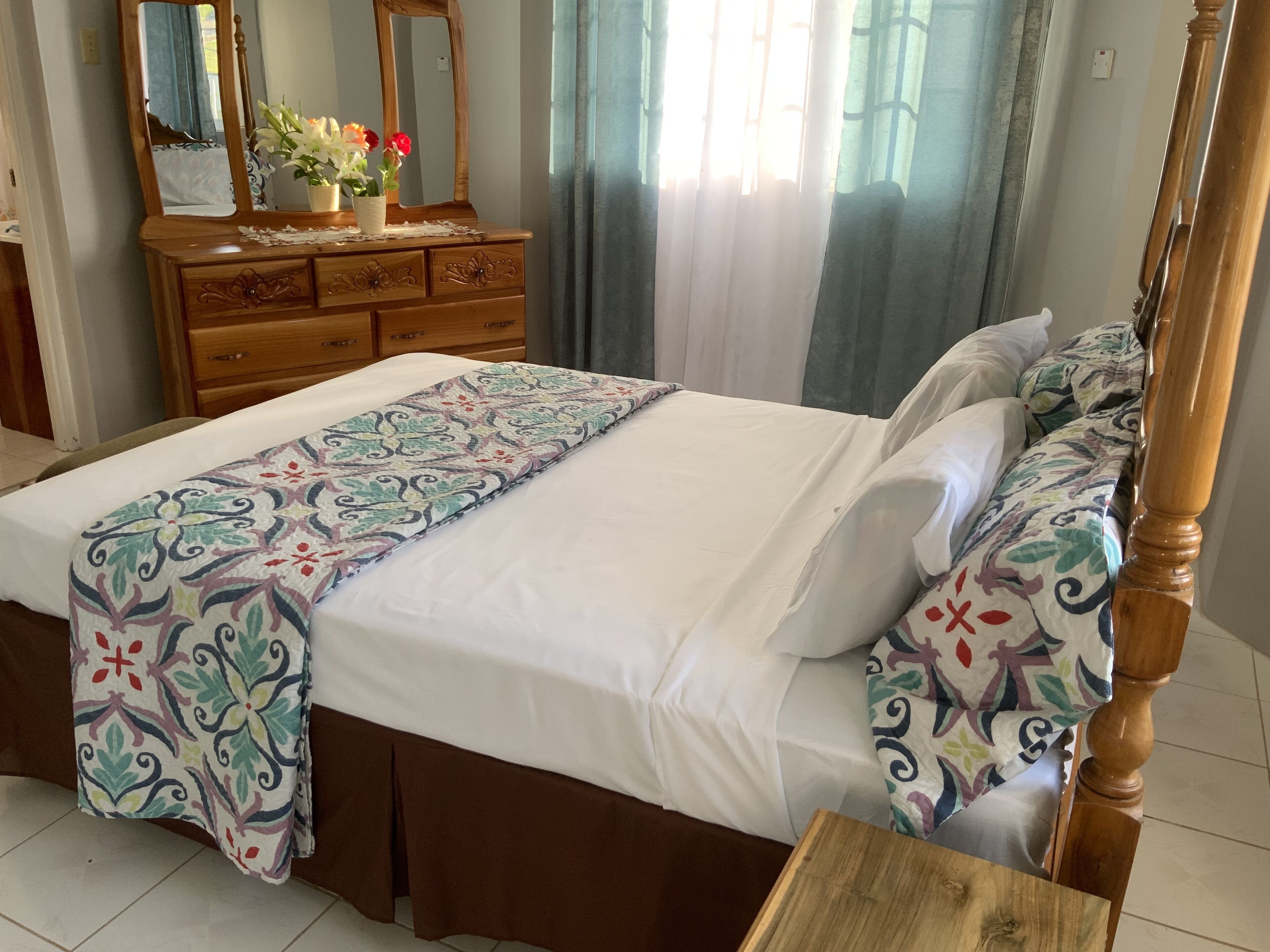 A neatly made bed with a patterned bedspread, a wooden dresser with a mirror, and a vase with flowers.