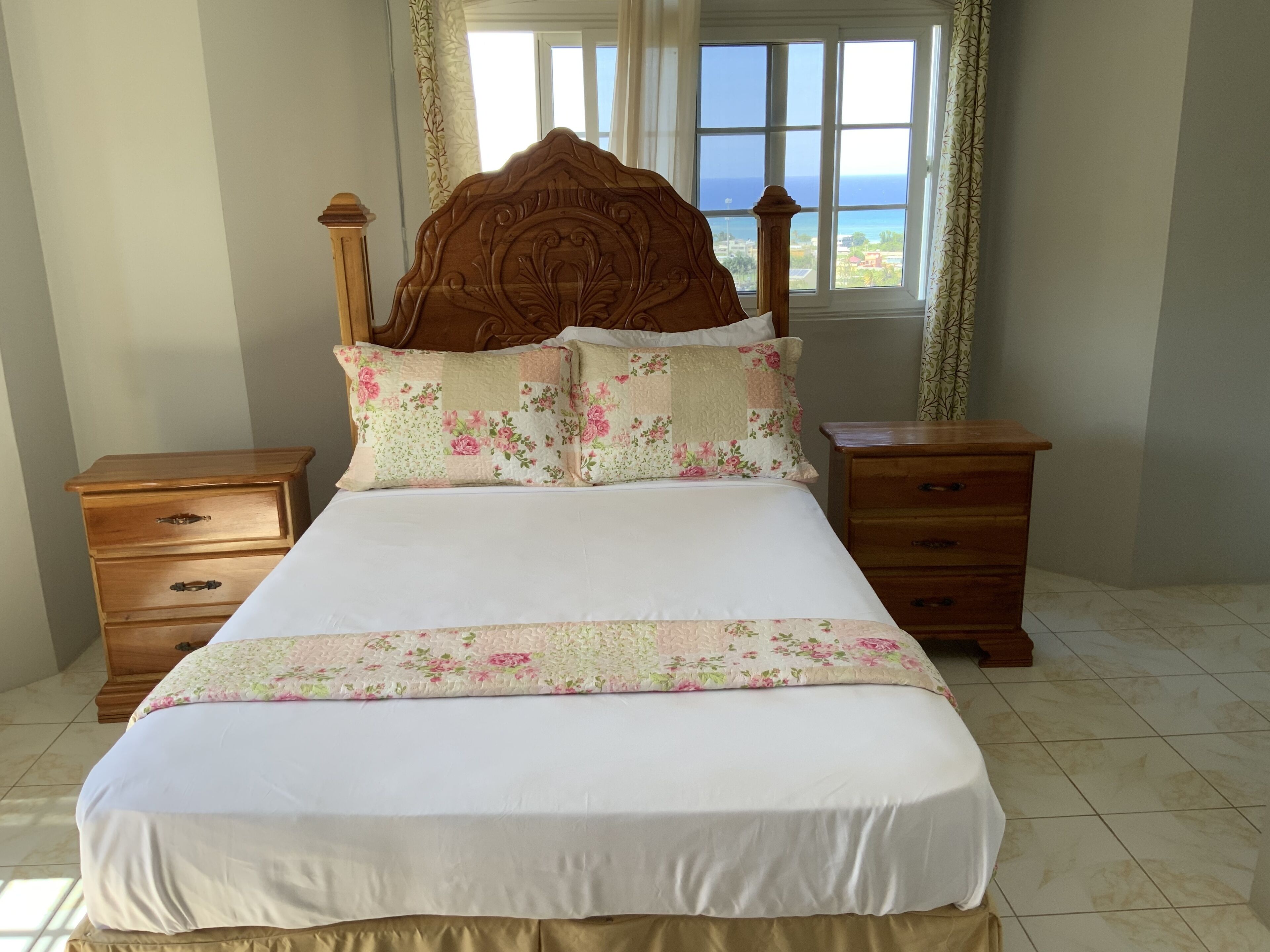 Una cama bien hecha con ropa de cama floral y un cabecero de madera, flanqueada por mesitas de noche de madera con cajones, en una habitación con un ventanal que ofrece vistas al mar.