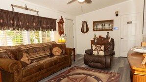 A living room with a brown leather sofa, a leather armchair, a wooden coffee table, a patterned rug, a wall clock, and a framed picture.