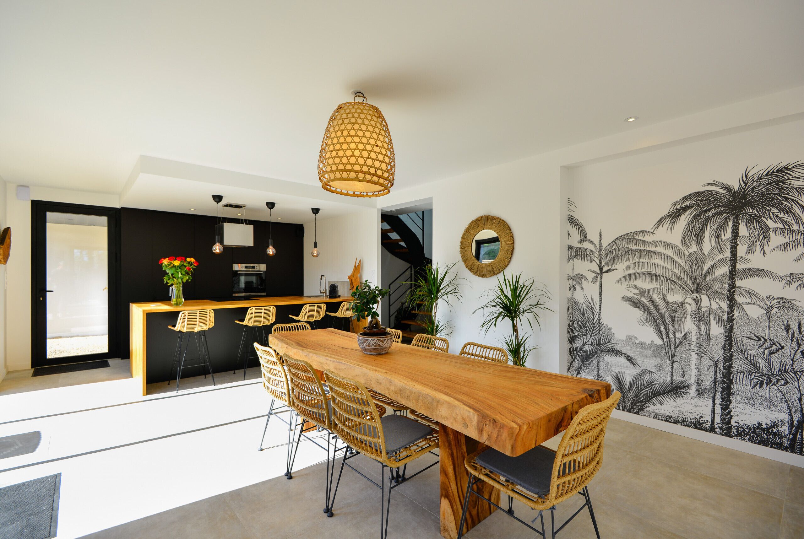 A modern dining area with a wooden table and wicker chairs, a kitchen in the background, and a tropical mural on the wall.