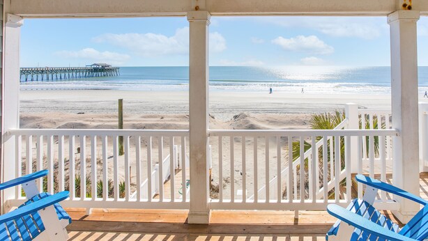 A beach view from a covered porch with white railings and blue chairs, overlooking the ocean and a pier.