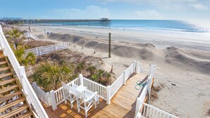 A beach view from a wooden deck with white railings, a pier in the background, and a sandy beach.