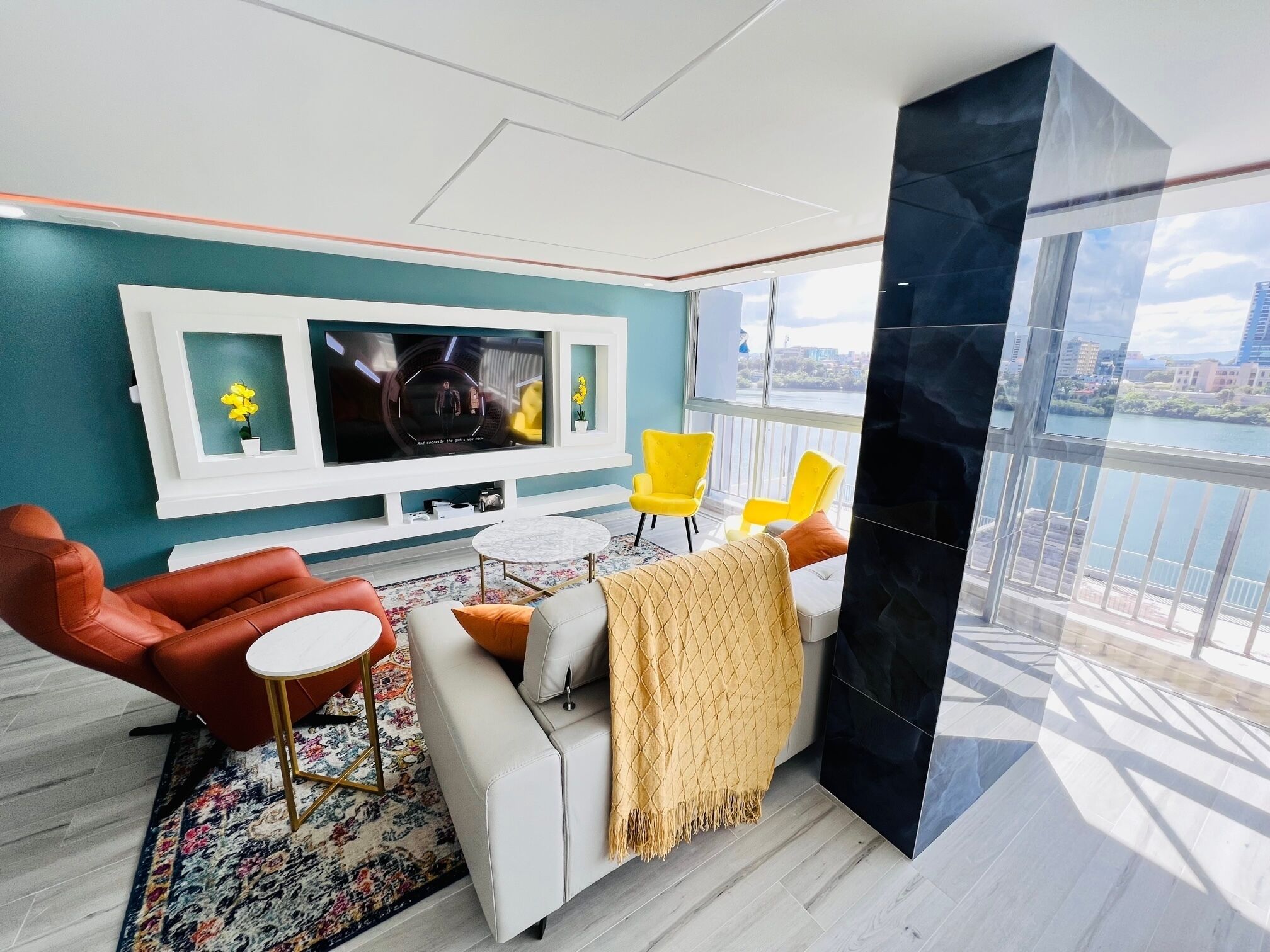 A modern living room with a large TV, two yellow chairs, a white sofa, and a view of a cityscape through large windows.