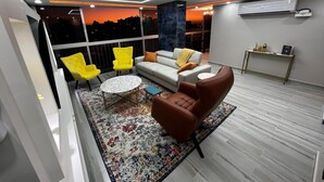 Un salon moderne avec un canapé, des fauteuils et une table basse. Une grande fenêtre offre une vue sur la ville.