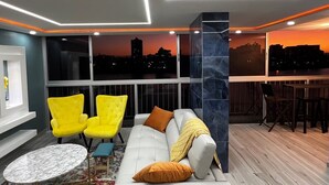 Un salon moderne avec un canapé, deux fauteuils jaunes, une table basse ronde et une vue sur le paysage urbain au coucher du soleil.