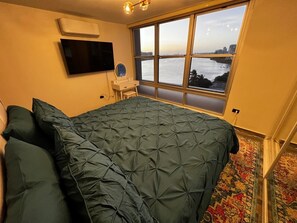 A bedroom with a large bed, a TV mounted on the wall, a window with a view of a body of water, and a small table with a mirror.