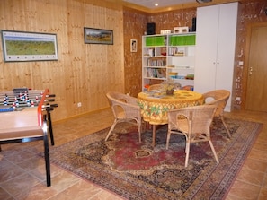 Een knus interieur met een tafelvoetbaltafel, rieten stoelen, een ronde tafel met een bloemenkleed en een boekenplank vol boeken en decoratieve objecten.
