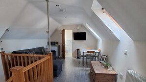Un loft espaiós amb un sostre alt, una escala de fusta, una zona de menjador amb taula i cadires, i un televisor fixat a la paret.