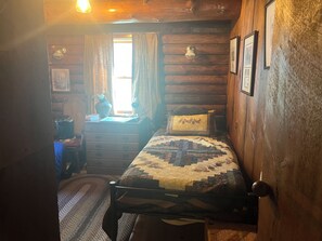 Una habitación tipo cabaña de troncos con una cama, un escritorio con lámpara y una ventana con cortinas.