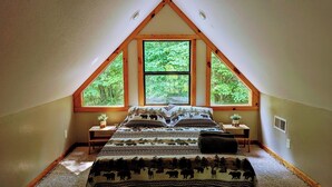 Une chambre avec un plafond en pente, une grande fenêtre donnant sur les arbres, un lit recouvert d’une couvre-lit à motifs, deux tables de chevet avec des lampes et une petite plante sur chaque table.