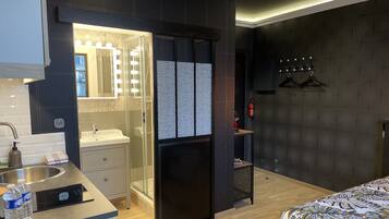 Ein modernes Hotelzimmer mit einem Badezimmer, das ein Waschbecken mit Spiegel und Beleuchtung, eine Spüle und eine Dusche bietet. Es gibt ein Bett mit einem gemusterten Überwurf und einen kleinen Holztisch mit einer Wasserflasche.
