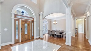 A spacious foyer with a wooden floor, a large white rug, a dining area with a chandelier, and a door with stained glass.