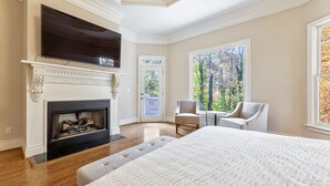 A bedroom with a fireplace, a TV mounted above it, a large window with a view of trees, and a bed with a white quilt.