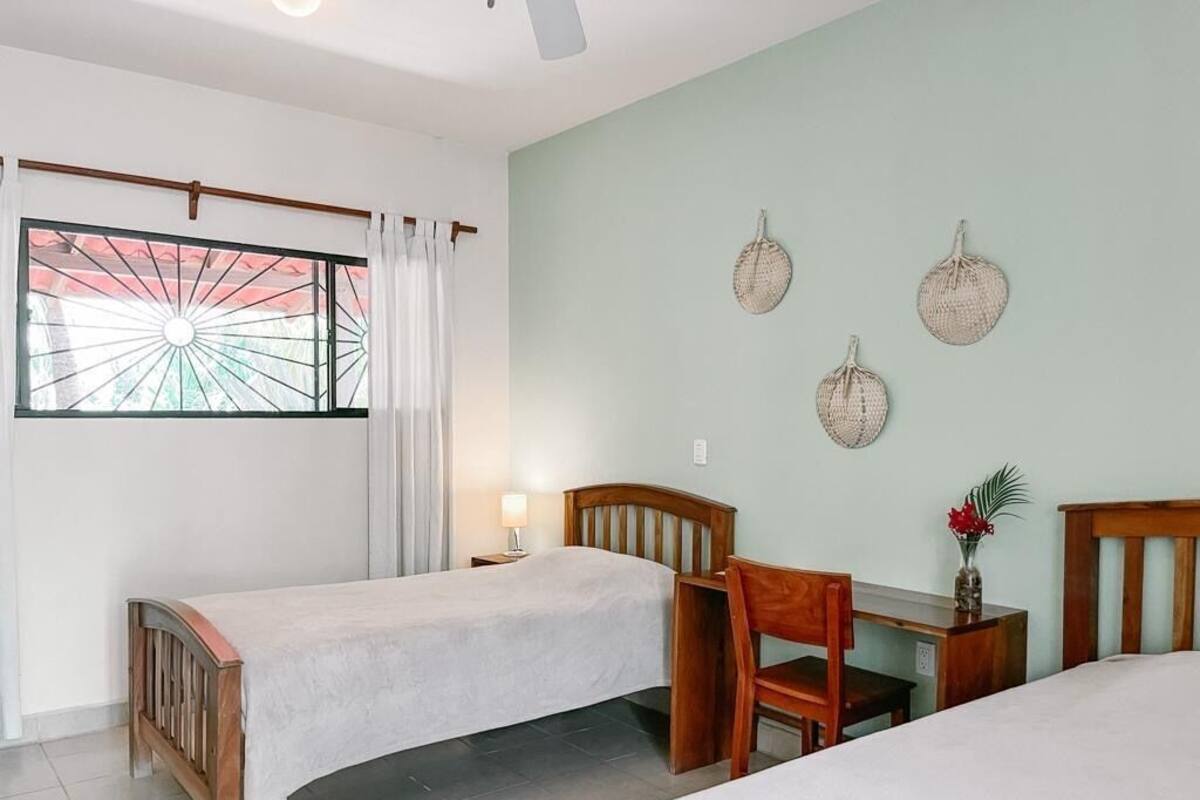 A room with two beds, a wooden chair, a small table, a window with curtains, and wall decorations.