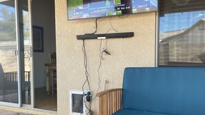 Un televisor fijado a la pared mostrando un partido de fútbol, una mesa de madera con un control remoto, una taza y un paño blanco doblado.