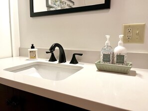 Un lavabo con grifo negro, un espejo encima y un jabonera verde con dos botellas de jabón de manos.