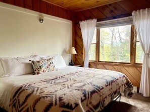 Un dormitorio de cabaña de madera con cama, cortinas blancas y ventana con vista a árboles.