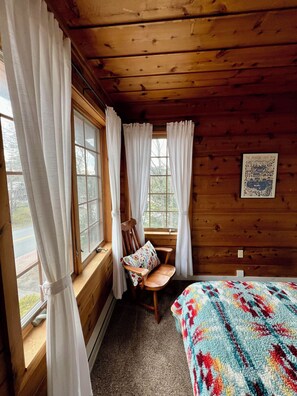 A cozy cabin room with wooden walls, a wooden bench with a cushion, a bed with a colorful blanket, and a window with white curtains. - Centrally Located Apartment (Lake Placid)