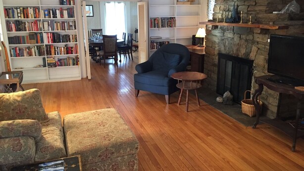 Ein gemütliches Wohnzimmer mit Kamin, blauem Sessel, gemustertem Sofa und einer Bücherwand voller Bücher.
